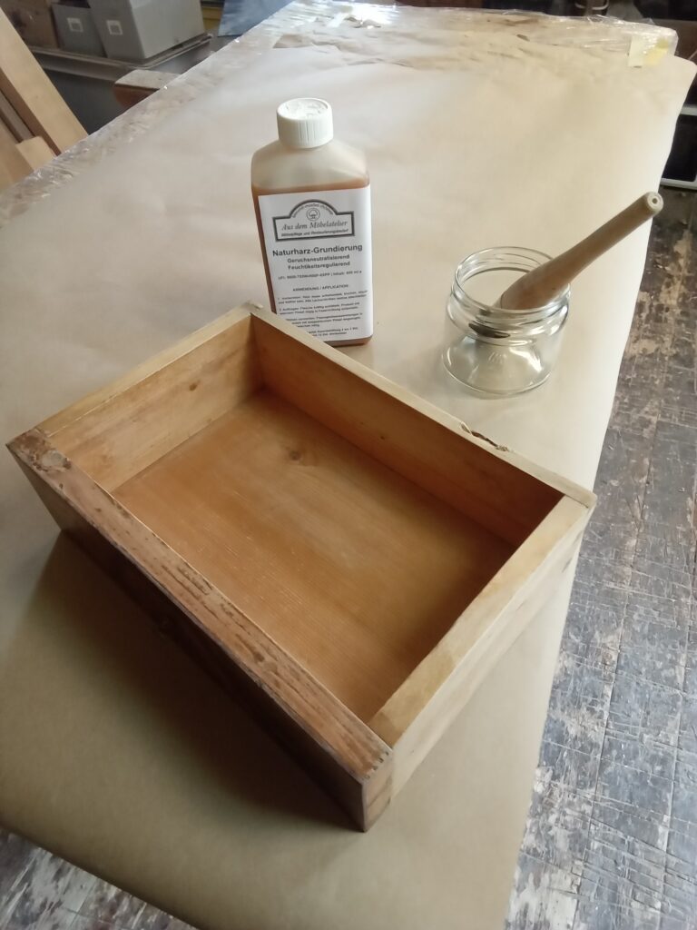 Unbehandeltes Massivholz einer geöffneten antiken Schublade neben einer Flasche der Naturharz-Grundierung zur Geruchsneutralisierung.